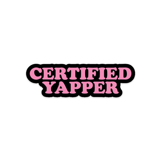 A rectangular sticker with a red background and a white horizontal stripe in the middle. In the center of the white stripe, black Devanagari text reads "घुमक्कड़" which translates to "Wanderer." The red background has faint travel-themed illustrations.